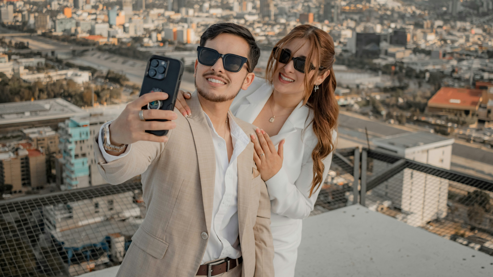 a well dressed young couple with sunglases are taking a selfie on a rooftop with city views